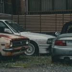 A rusted vintage car alongside modern vehicles in a gritty urban backdrop, highlighting contrast.